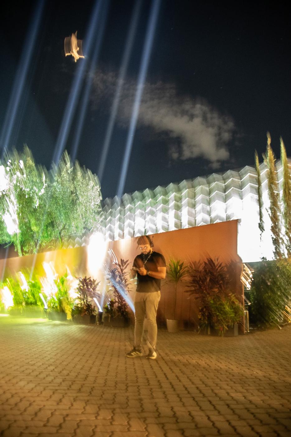 Membro da equipe em captação noturna com drone no céu e fachada iluminada